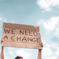 Woman holding a banner of 'We Need A Change'. Young woman with poster in front of people protesting about climate changing on the street. Meeting about problem in ecology, environment, global warming, industrial influence, climate emergency.