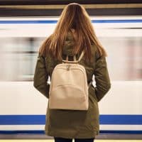 Back view of unrecognizable female passenger with rucksack standing with hands in pockets on underground station with blurred moving train