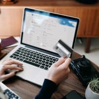 Over the shoulder view of young Asian woman shopping online for flight tickets on airline website with laptop, entering credit card details to make mobile payment at home. Camera and passport on the table. Travel planning. Booking a holiday online