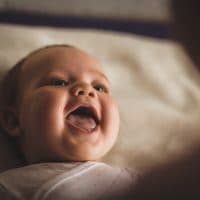 High angle view of cheerful newborn baby girl lying down on the bed and laughing while her mother, that is blurred in foreground, is playing with her.