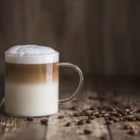 Cafe latte macchiato layered coffee in a see through glass coffee cup. The cup is on a wooden background with coffee beans on the table next to the cup.