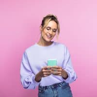 Happy beautiful woman using smart phone while standing against pink background.