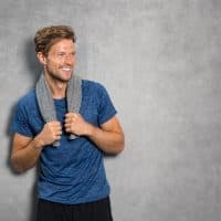 Portrait of a smiling fitness man with towel on shoulders looking away. Happy young man relaxing after training and leaning on grey wall. Sporty active guy resting after workout with copy space.