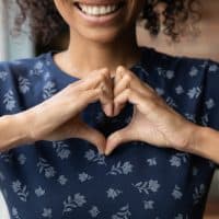 Crop close up of happy African American woman feel grateful thankful show heart sign spread love and care. Smiling biracial female volunteer make hand gesture support ill sick people patients.