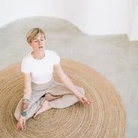 Beautiful authentic woman with tattoos and short blond hair is meditating sitting in lotus position on wicker carpet in minimalist home interior. She is wearing a light-colored casual clothing. Concept of relaxation exercises