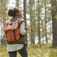 Back view portrait of young African-American woman with backpack enjoying hiking in forest lit by sunlight, copy space