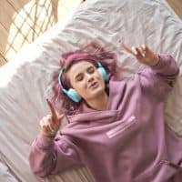 Happy funny teen girl with pink hair wear headphones lying in comfortable bed listening new pop music enjoying singing song with eyes closed relaxing in cozy bedroom at home. Top view from above.