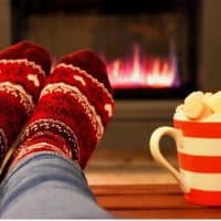 Stock photo of frothy hot chocolate drink in mug with gas fire in background and feet on the coffee table at Christmas . This man is wearing socks to warm up in winter weather. The hot chocolate is topped with delicious pink and white marshmallow sweets