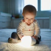 Adorable baby girl playing with bedside lamp in nursery. Happy kid sitting on bed with nightlight. Little child at home in the evening before sleep
