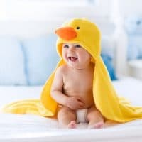 Happy laughing baby wearing yellow hooded duck towel sitting on parents bed after bath or shower. Clean dry child in bedroom. Bathing and washing of little kids. Children hygiene. Textile for infants.