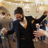 Man at a wedding dressed in a suit and sunglasses dancing while other guests stand around him. He is holding a glass of wine and laughing.