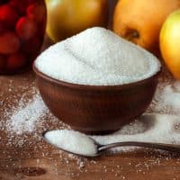 White sugar in a ceramic bowl of red clay and in a teaspoon on a wooden table with apples.