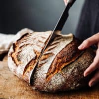 Close-up of woman cutting sourdough bread on board. Midsection of female is with knife and baked bread. She is in kitchen.