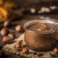 Front view of a crystal jar full of chocolate and hazelnut spread surrounded by some hazelnuts on a rustic wooden table. Selective focus is on the jar and on the defocused background are some croissants and chocolate bars. Predominant color is brown. Low key DSLR photo taken with Canon EOS 6D Mark II and Canon EF 24-105 mm f/4L