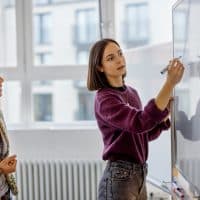 Businesswoman brainstorming ideas on whiteboard with colleague. Female business partners having brainstorming session in startup meeting room.