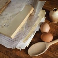 Old recipe books and hand-written recipes on a kitchen worktop, along with a pie ceramic funnel, eggs and a wooden spoon.