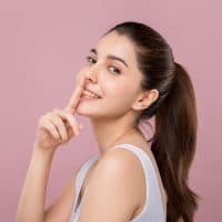 Young beautiful Caucasian woman with smiley face touching her nose, surgery nose job concept, isolated on pink background.