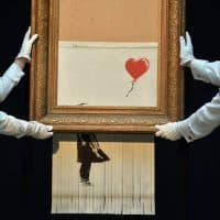 Sotheby's employees pose with the newly completed work by artist Banksy entitled "Love is in the Bin", a work that was created when the painting "Girl with Balloon" was passed through a shredder in a surprise intervention by the artist, at Sotheby's auction house in London on October 12, 2018, following the work's sale. The buyer of a work by street artist Banksy that was partially destroyed moments after it sold has gone through with the purchase, Sotheby's auction house said on October 11, 2018. The painting "Girl with Balloon" was passed through a shredder hidden in the frame just after it went under the hammer last week for £1,042,000 ($1.4 million, 1.2 million euros). The modified version has now been certified by Banksy's authentication body Pest Control as a new piece of work in its own right, entitled "Love is in the Bin". (Photo by Ben STANSALL / AFP) / RESTRICTED TO EDITORIAL USE - MANDATORY MENTION OF THE ARTIST UPON PUBLICATION - TO ILLUSTRATE THE EVENT AS SPECIFIED IN THE CAPTION