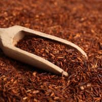 Heap of dry rooibos tea leaves with wooden scoop, closeup view