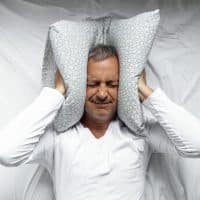 One mature man trying to sleep covering his ears to avoid neighbour noise at home or hotel during the day.