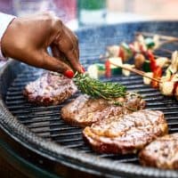 Steaks being seasoned with herbs as they cook on bbq grill with vegetable kebabs, mediterranean food, healthy lifestyle, food and drink