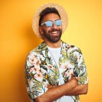 Indian man on vacation wearing floral shirt hat sunglasses over isolated yellow background happy face smiling with crossed arms looking at the camera. Positive person.