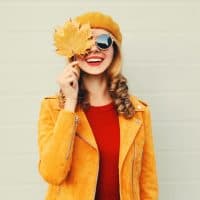 Autumn mood! happy smiling woman holding in her hands yellow maple leaves hiding her eye over gray wall background