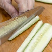 A chef julienning a cucumber into long thin strips on a wooden cutting board while wearing plastic gloves.