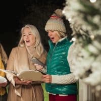 Group of three women dressed warmly standing at a front door holding books as they Christmas carol.