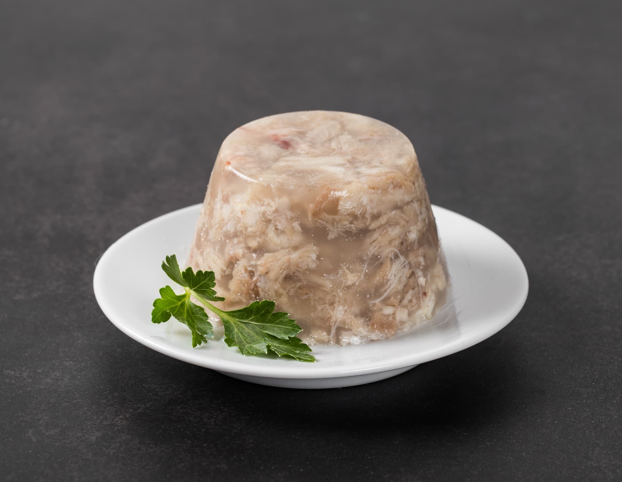 Traditional Russian festive cold appetizer. Pork jelly with pork slices and parsley on a plate on a dark gray background. Dish of thickened to a jelly-like mass from cooling meat broth with pieces of meat