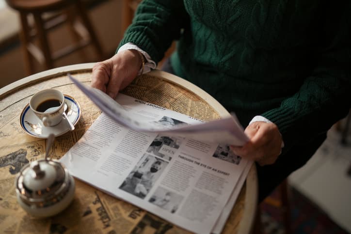 Midsection of older woman with newspaper sitting by table in coffee shop and having morning coffee, cut out.
