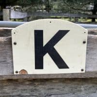 A large placard with the letter K written on it in black is attached to a wooden wall. It is used in dressage.part of it is damaged.