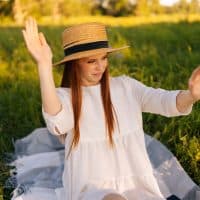 Portrait of dissatisfied redhead pretty woman in straw hat and white dress chasing insects away from herself sitting on field with green grass, on background of soft sunlight in summer sunny day.