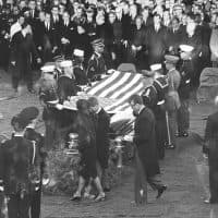 At Arlington National Cemetary, Jacqueline Kennedy (1929 - 1994), accompanied by her brothers-in-law Robert F. Kennedy (1925 - 1968) and Ted Kennedy (1932 - 2009), approach the gravesite of assassinated President John F. Kennedy's receive the flag being folded by a group of servicemen representing all branches of the US armed services, Virginia, November 25, 1963. Visible in the background is US President Lyndon B. Johnson (1908 - 1973) among various international dignitaries. (Photo by Abbie Rowe/PhotoQuest/Getty Images)