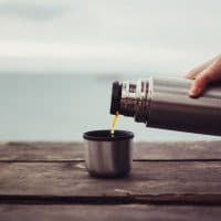 Unrecognizable traveler pouring tea to cup from thermos on wooden table on background of sea. Theme tourism and travel